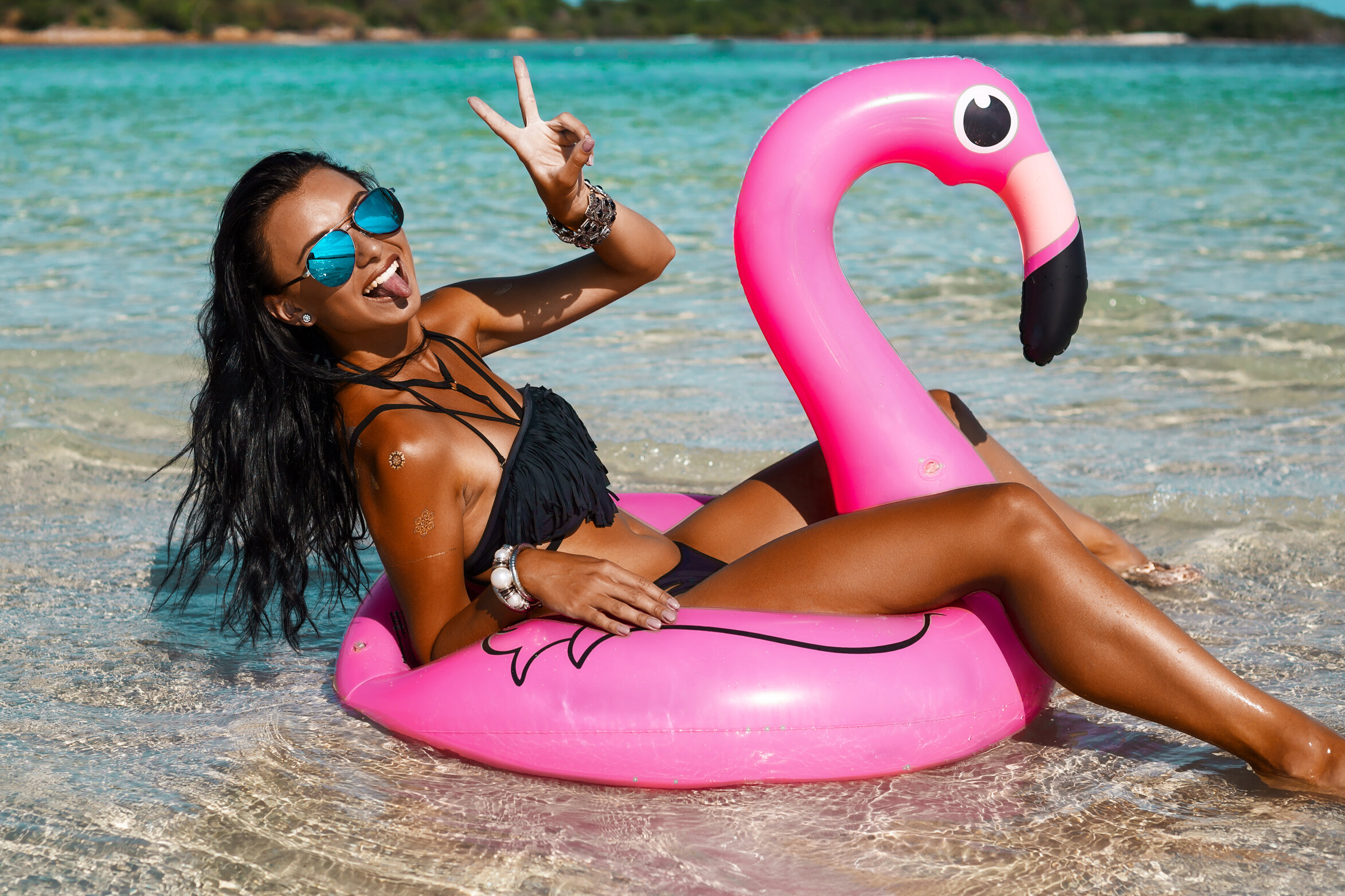 A beautiful sexy amazing young woman on the beach sits on an inflatable pink flamingo and laughs, has a great time, tanned perfect body, long hair, black bikini, fashion accessories, low key photo